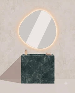 A modern bathroom vanity setup with a dark green marble sink block and a large organic-shaped wall mirror featuring a warm glowing LED light border, set against a textured light beige wall.