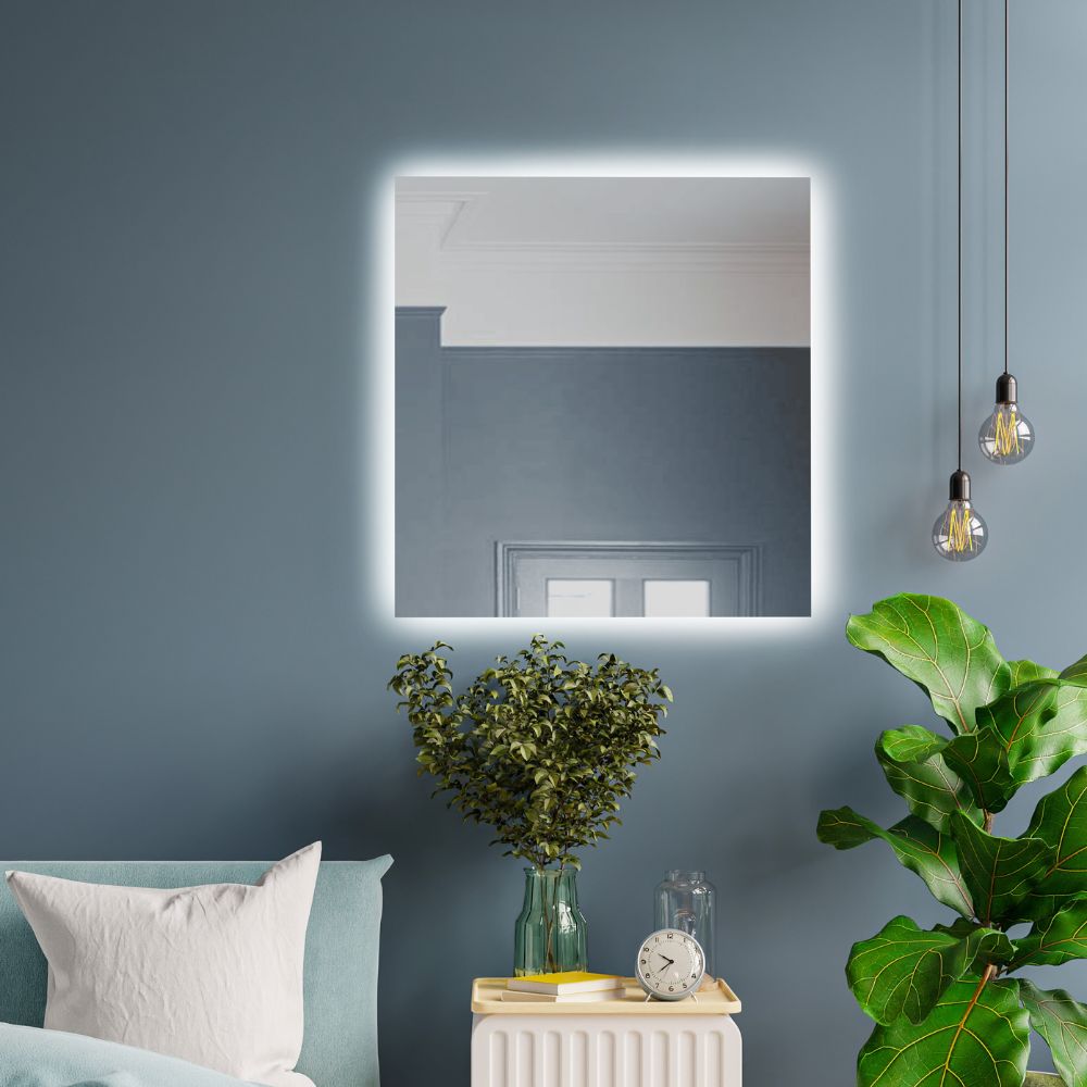 A square LED backlit mirror with rear lighting mounted on a blue wall, surrounded by a dark room with hanging lights and plants.