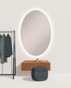 An oval backlit vanity mirror mounted on a light beige wall above a wooden console table, with a dark woven pouf in front and a clothing rack with dark garments to the left.