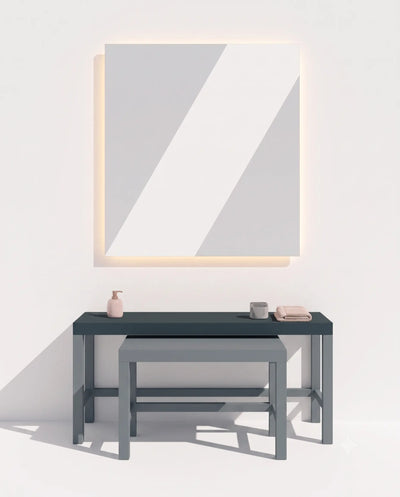 A minimalist bathroom scene with a large rectangular wall mirror featuring a glowing warm light border, above a dark gray console table with a matching stool and small toiletry items.